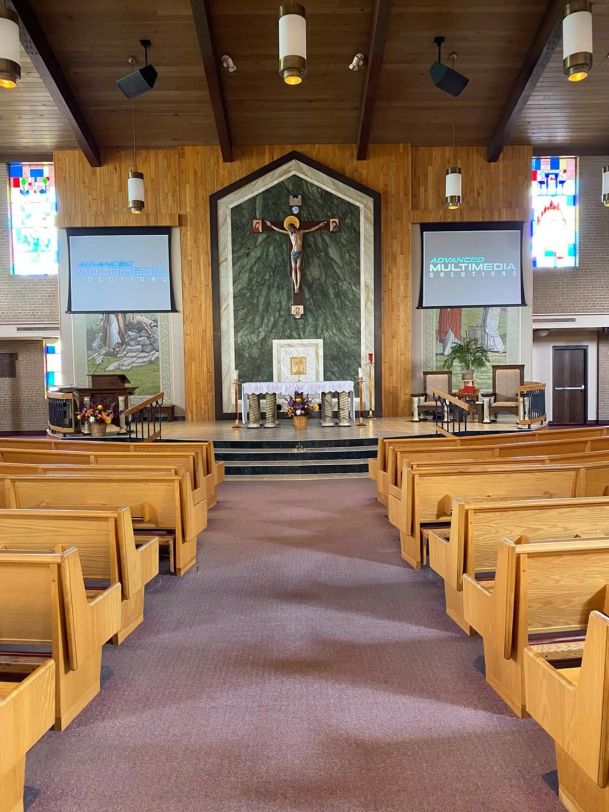 Church interior with wooden pews facing an altar, crucifix, and dual projection screens