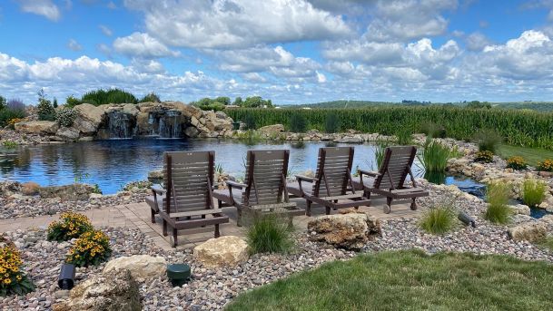Four lounge chairs facing a backyard pond with a stone waterfall next to a field
