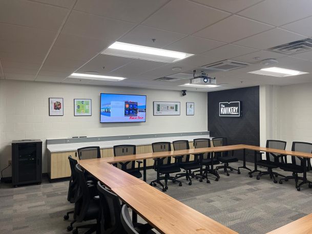 Conference room with U-shaped wooden tables, office chairs, and a wall display screen