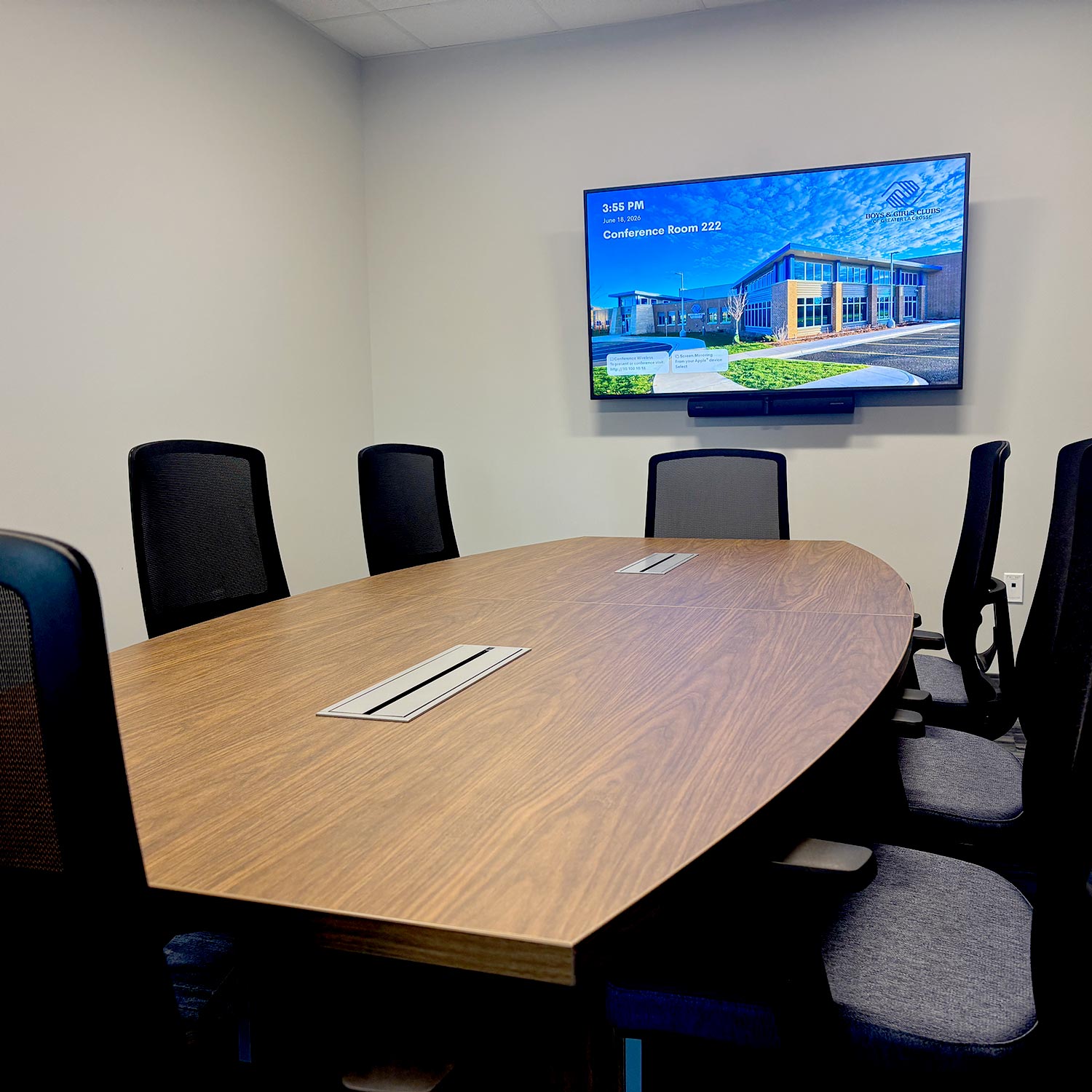Conference room with a wooden table, black office chairs, and a wall-mounted digital display