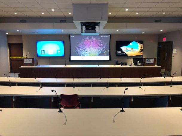 Large presentation hall with rows of desks equipped with microphones facing multiple screens
