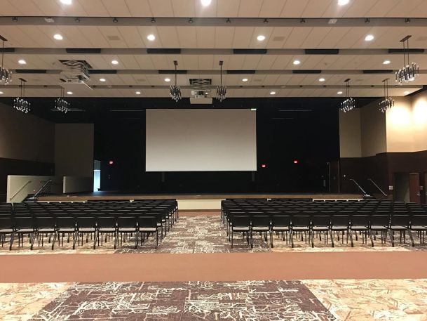 Spacious event hall with rows of black chairs arranged in front of a projection screen